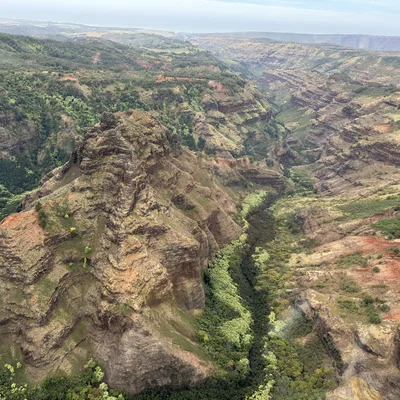 An aerial view captures a vast canyon landscape featuring a prominent, rugged mountain peak in the foreground and a deep valley filled with green vegetation. The surrounding terrain consists of layered rock formations in shades of brown, red, and green under a slightly overcast sky.