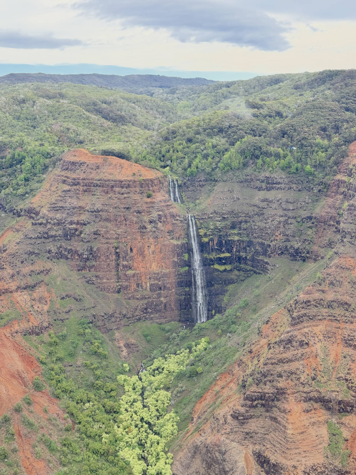 A scenic view of a tall, multi-tiered waterfall cascading down a steep, reddish-brown canyon wall surrounded by lush green vegetation. The landscape features rugged, forested hills under a cloudy sky.