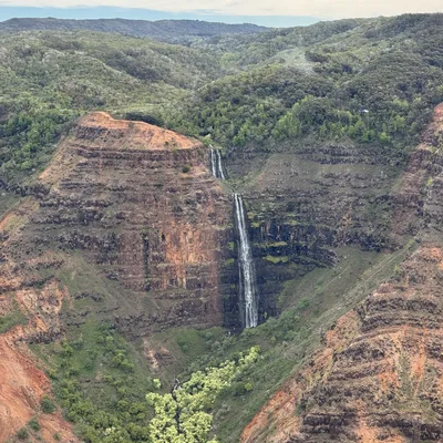 A scenic view of a tall, multi-tiered waterfall cascading down a steep, reddish-brown canyon wall surrounded by lush green vegetation. The landscape features rugged, forested hills under a cloudy sky.