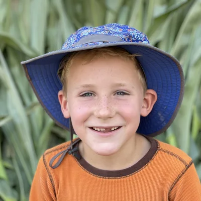 A young boy with freckles and a missing front tooth smiles at the camera while wearing a blue sun hat and an orange shirt. He is positioned outdoors in front of a dense background of green corn stalks.