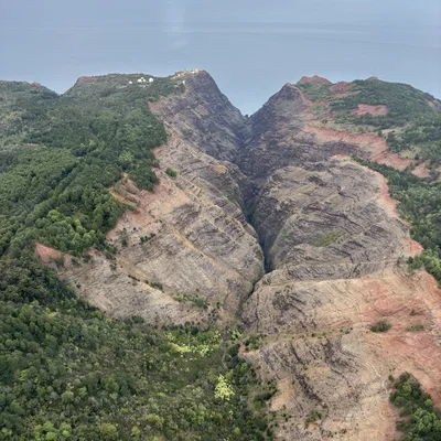 An aerial view captures a deep, rugged canyon with steep, layered rock walls and green forested ridges leading toward the ocean in the background. White dome-shaped structures are visible on a distant clifftop overlooking the sea.