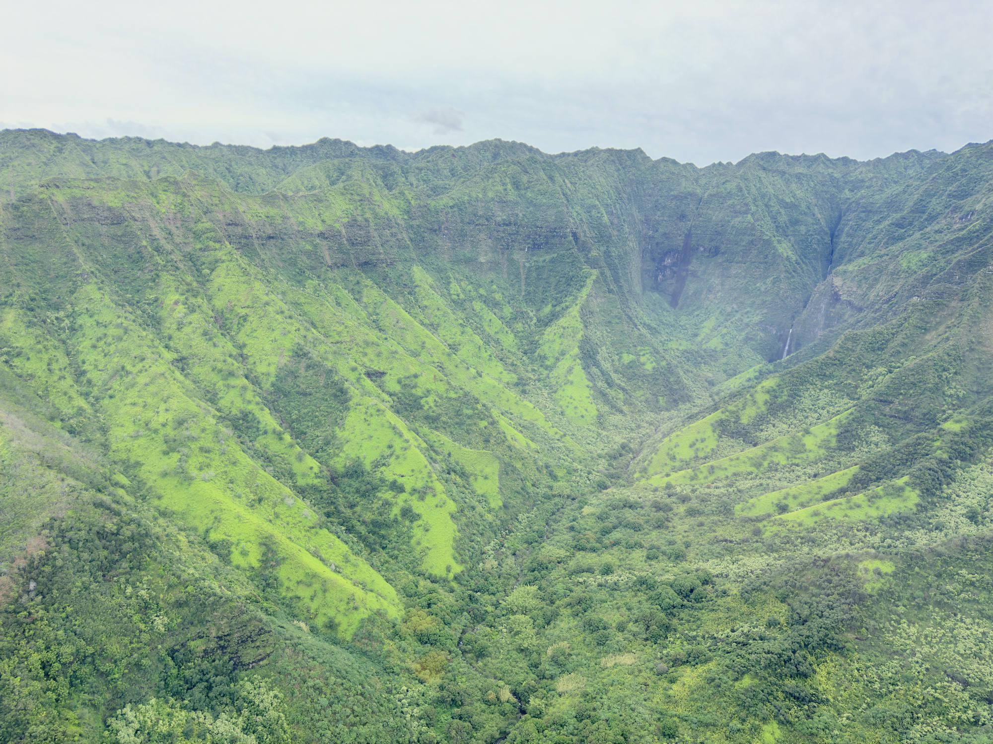 An aerial view captures a lush, green mountain valley with steep, ridged slopes and a distant waterfall under a cloudy sky.