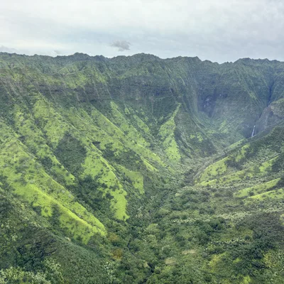 An aerial view captures a lush, green mountain valley with steep, ridged slopes and a distant waterfall under a cloudy sky.