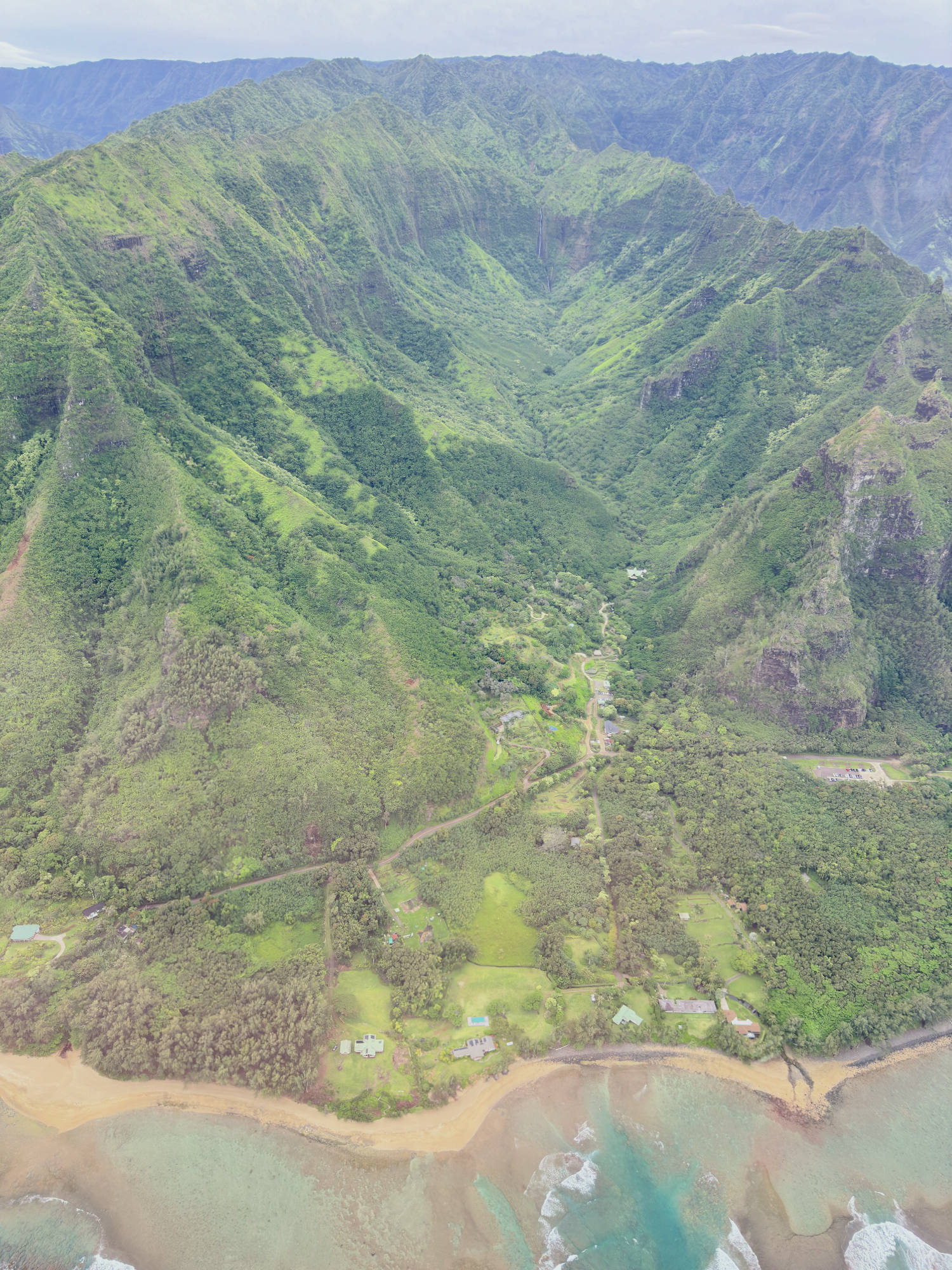 An aerial photograph captures a lush, green mountain valley meeting a sandy beach and turquoise ocean. Scattered houses and winding roads are visible nestled in the forested landscape at the base of the steep, verdant ridges.