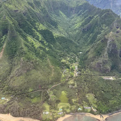 An aerial photograph captures a lush, green mountain valley meeting a sandy beach and turquoise ocean. Scattered houses and winding roads are visible nestled in the forested landscape at the base of the steep, verdant ridges.