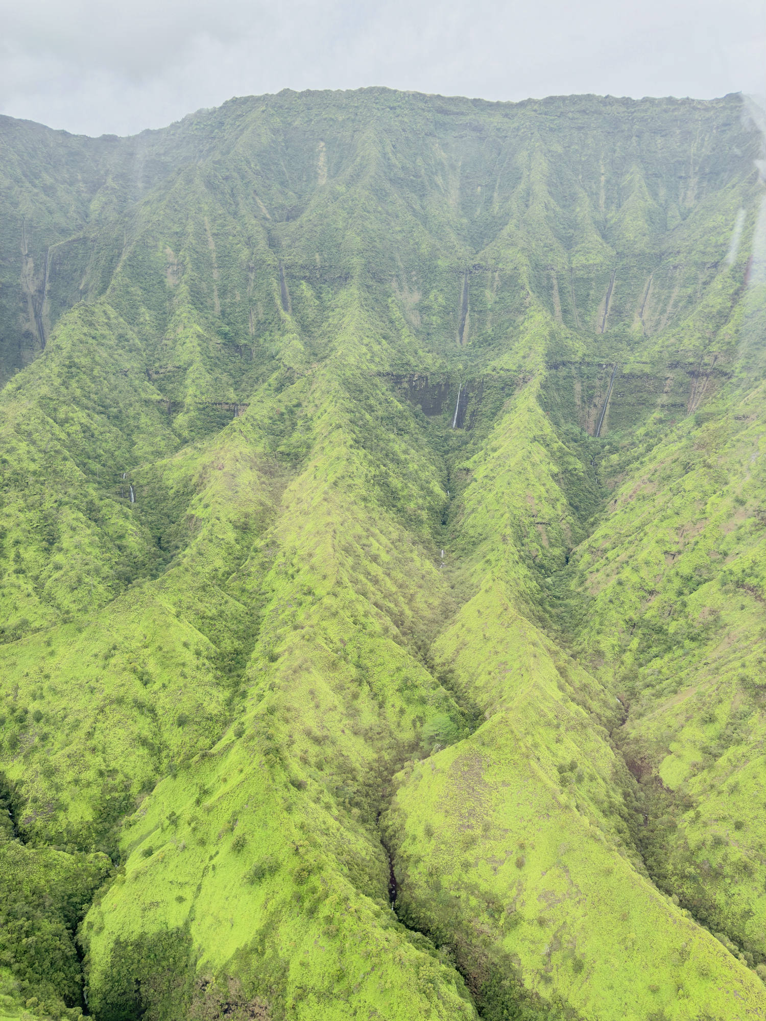 An aerial view captures a vast, lush green mountain range with steep, ridged slopes under a cloudy sky. Several thin waterfalls cascade down the deep crevices of the verdant, forested valleys.