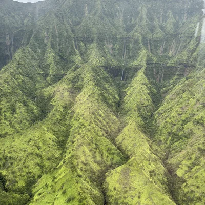 An aerial view captures a vast, lush green mountain range with steep, ridged slopes under a cloudy sky. Several thin waterfalls cascade down the deep crevices of the verdant, forested valleys.
