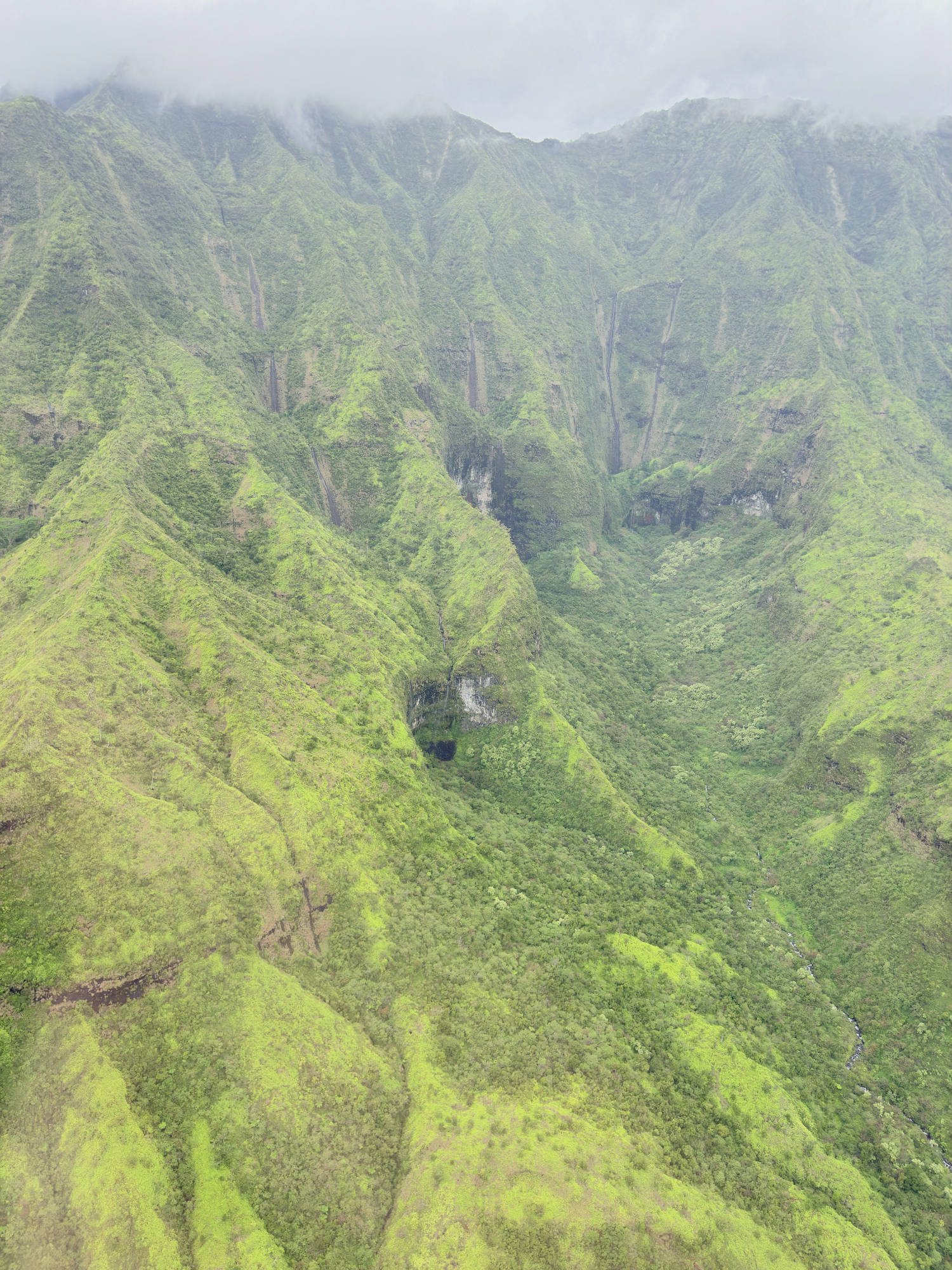 An aerial view captures a lush, green mountain valley with steep ridges and several thin waterfalls cascading down the slopes. Low-hanging clouds and mist shroud the peaks of the mountains in the background.