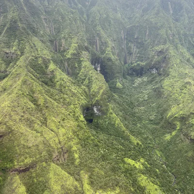 An aerial view captures a lush, green mountain valley with steep ridges and several thin waterfalls cascading down the slopes. Low-hanging clouds and mist shroud the peaks of the mountains in the background.