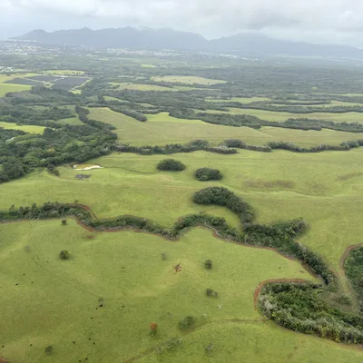 An aerial view captures a vast landscape of rolling green fields, pastures, and patches of forest under a cloudy sky. In the distance, mountains rise behind a coastal town, while a winding dirt path and a small pond are visible in the foreground.
