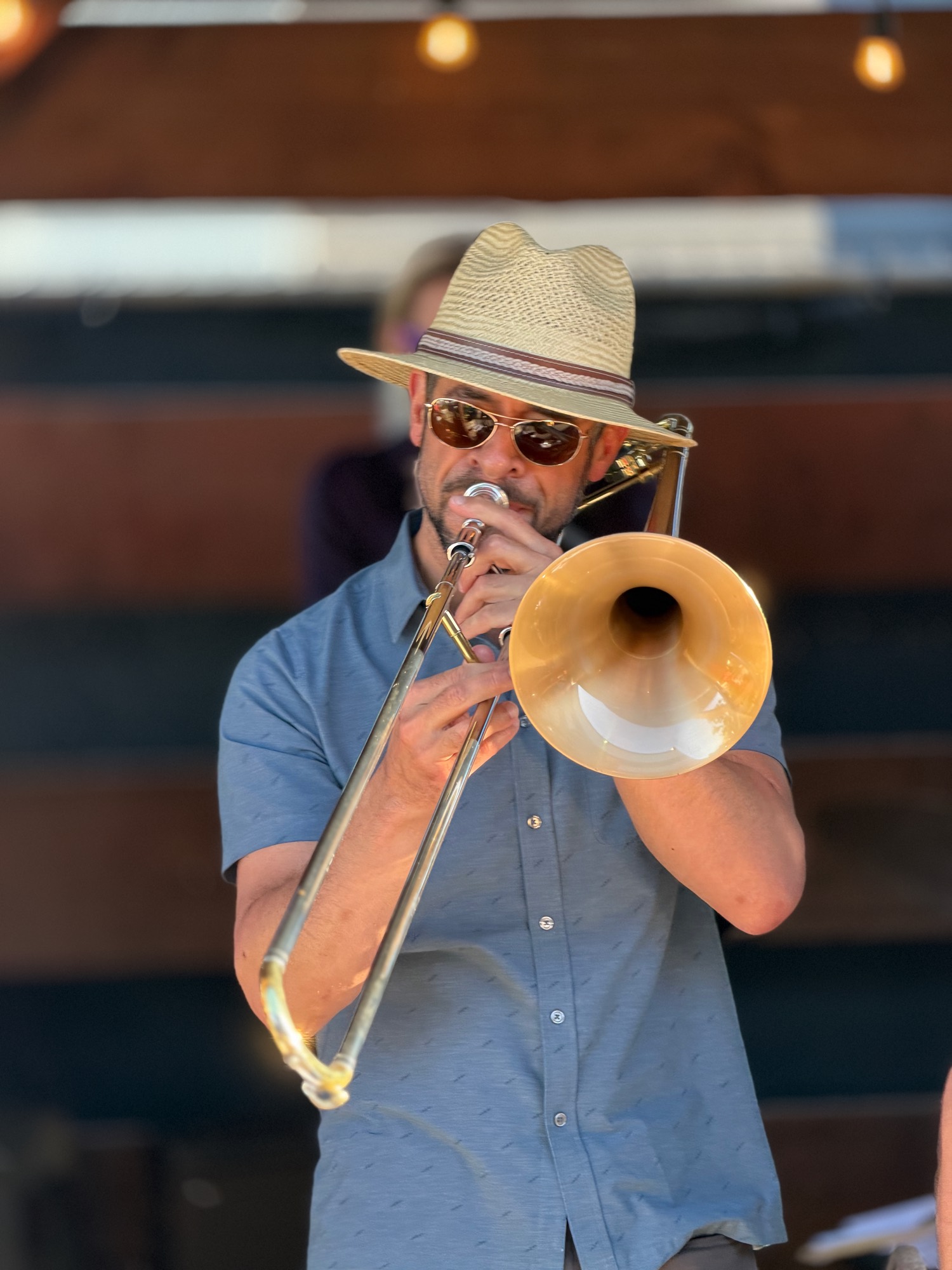 A man wearing a straw hat, sunglasses, and a blue button-down shirt plays a brass trombone. He is performing on an outdoor stage with another musician blurred in the background.
