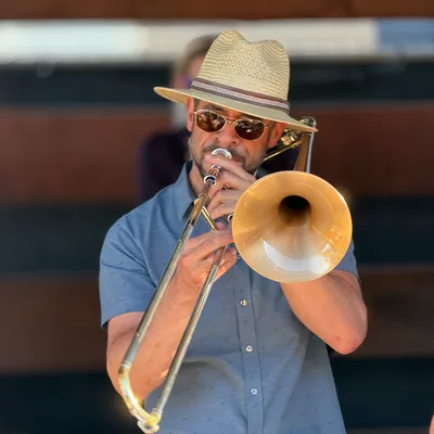 A man wearing a straw hat, sunglasses, and a blue button-down shirt plays a brass trombone. He is performing on an outdoor stage with another musician blurred in the background.