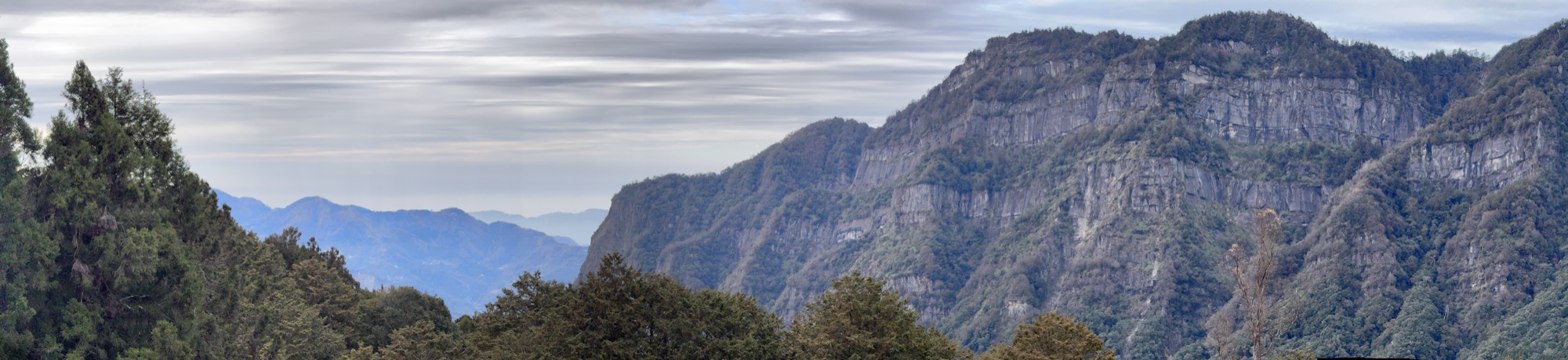 This panoramic photograph captures a vast mountain range under a cloudy, overcast sky, with steep, forested cliffs dominating the right side of the frame. In the foreground, dense green treetops line the ridge, while distant blue-toned mountains fade into the hazy background on the left.