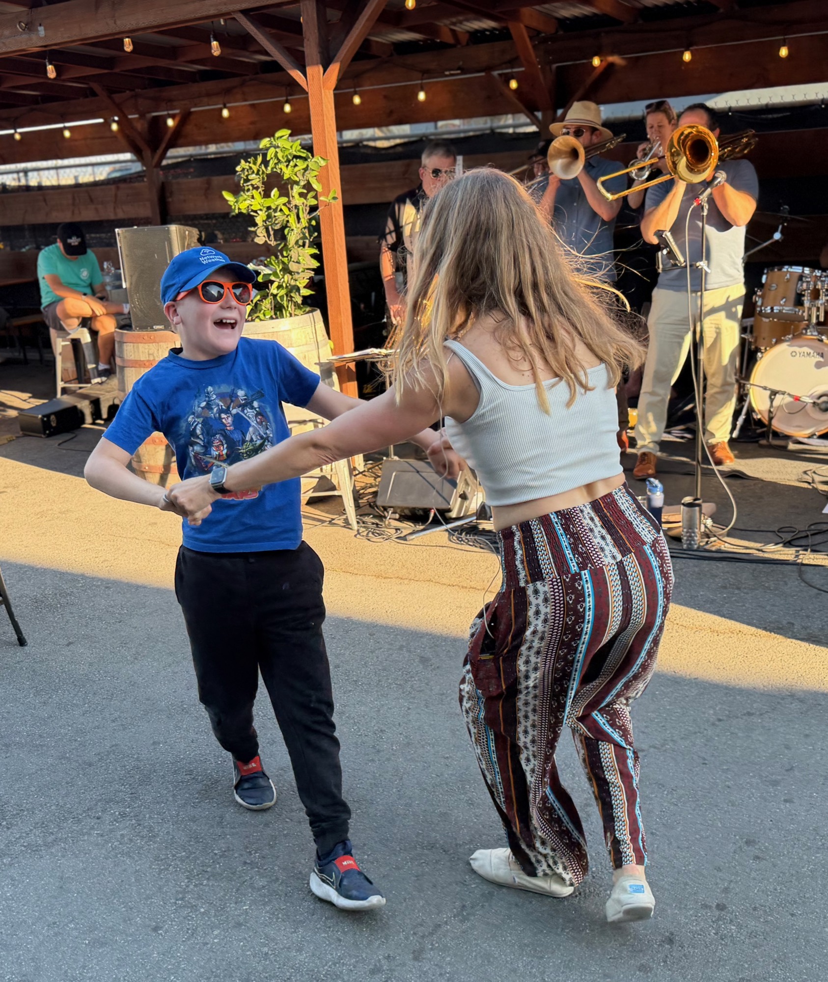 A young boy wearing sunglasses and a blue cap dances with a woman in patterned pants on an outdoor patio. In the background, a live brass band performs under a wooden pavilion structure.