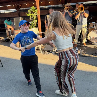A young boy wearing sunglasses and a blue cap dances with a woman in patterned pants on an outdoor patio. In the background, a live brass band performs under a wooden pavilion structure.