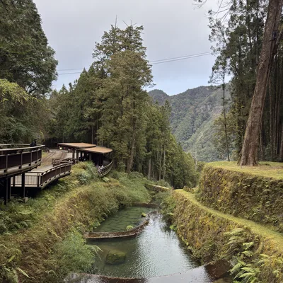 A tiered, stone-lined stream flows through a lush, forested mountain valley under an overcast sky. On the left, a wooden boardwalk and a covered train platform sit nestled among the trees, with distant mountains visible in the background.