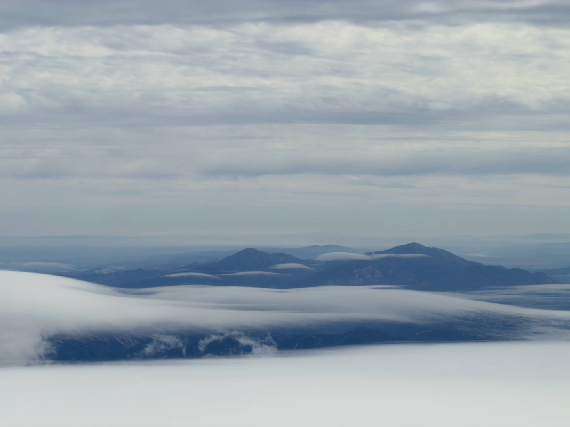 This landscape photograph captures dark blue mountain peaks rising above a thick layer of low-lying white clouds under a grey, overcast sky.