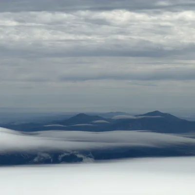 This landscape photograph captures dark blue mountain peaks rising above a thick layer of low-lying white clouds under a grey, overcast sky.