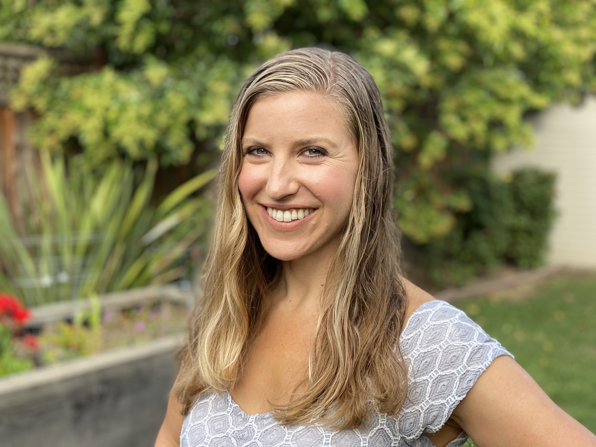 A smiling woman with long blonde hair poses for a portrait outdoors in a backyard setting. She is wearing a light-colored patterned top, with green foliage and a garden bed visible in the blurred background.