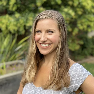 A smiling woman with long blonde hair poses for a portrait outdoors in a backyard setting. She is wearing a light-colored patterned top, with green foliage and a garden bed visible in the blurred background.