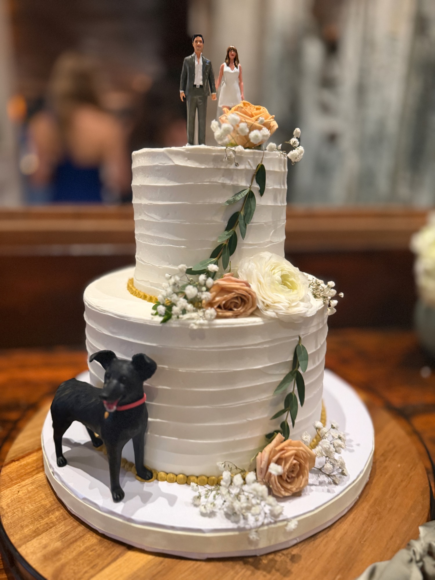 A two-tiered white wedding cake decorated with peach and white flowers, green leaves, and a black dog figurine sits on a wooden table indoors. The cake is topped with a custom figurine of a bride and groom.
