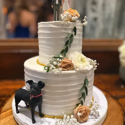 A two-tiered white wedding cake decorated with peach and white flowers, green leaves, and a black dog figurine sits on a wooden table indoors. The cake is topped with a custom figurine of a bride and groom.