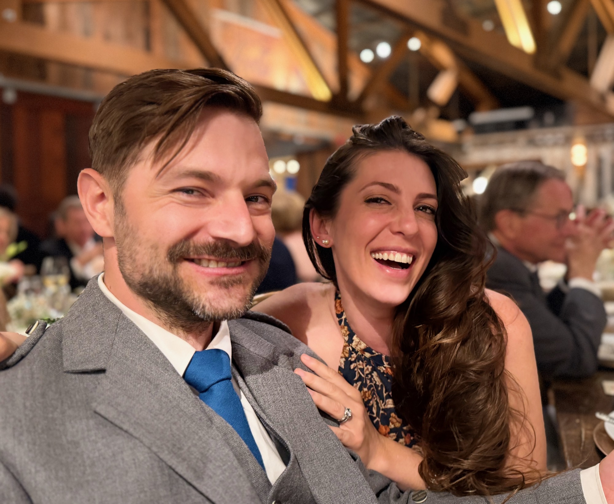 A smiling man in a grey suit and blue tie poses for a close-up photo with a laughing woman who has her hand on his shoulder. They are seated at a table in a dimly lit, rustic indoor venue with other guests visible in the background.