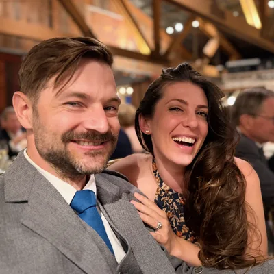 A smiling man in a grey suit and blue tie poses for a close-up photo with a laughing woman who has her hand on his shoulder. They are seated at a table in a dimly lit, rustic indoor venue with other guests visible in the background.