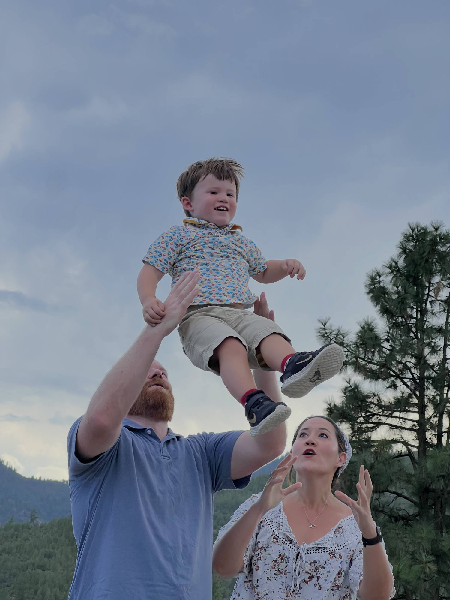 A man tosses a smiling young boy into the air while a woman watches with open arms in an outdoor setting with pine trees and mountains in the background.