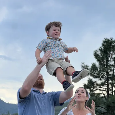 A man tosses a smiling young boy into the air while a woman watches with open arms in an outdoor setting with pine trees and mountains in the background.