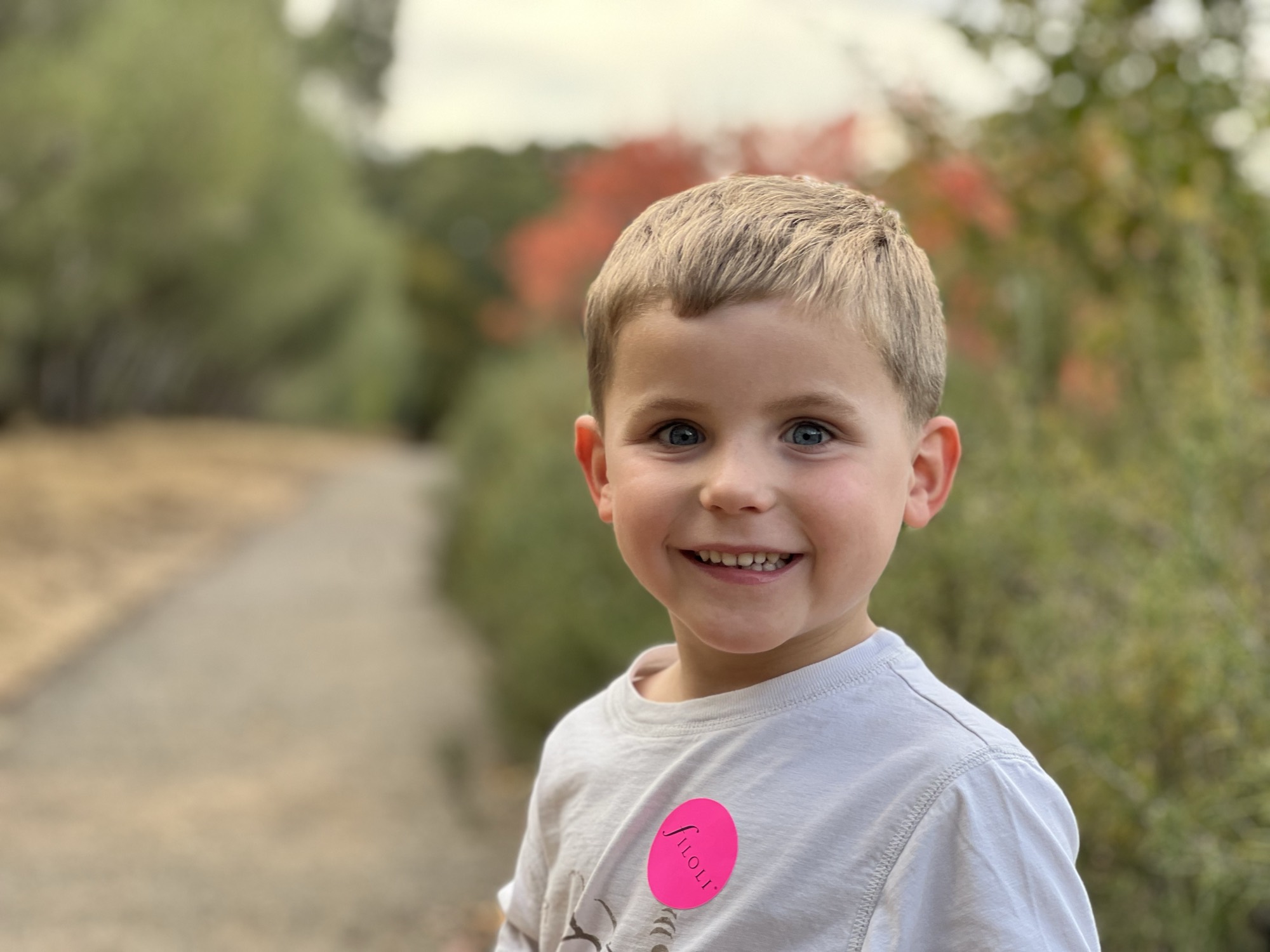 A close-up portrait of a smiling young boy with blue eyes wearing a white t-shirt with a pink "Filoli" sticker. He is standing outdoors on a dirt path with blurred trees and foliage in the background.