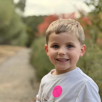 A close-up portrait of a smiling young boy with blue eyes wearing a white t-shirt with a pink "Filoli" sticker. He is standing outdoors on a dirt path with blurred trees and foliage in the background.