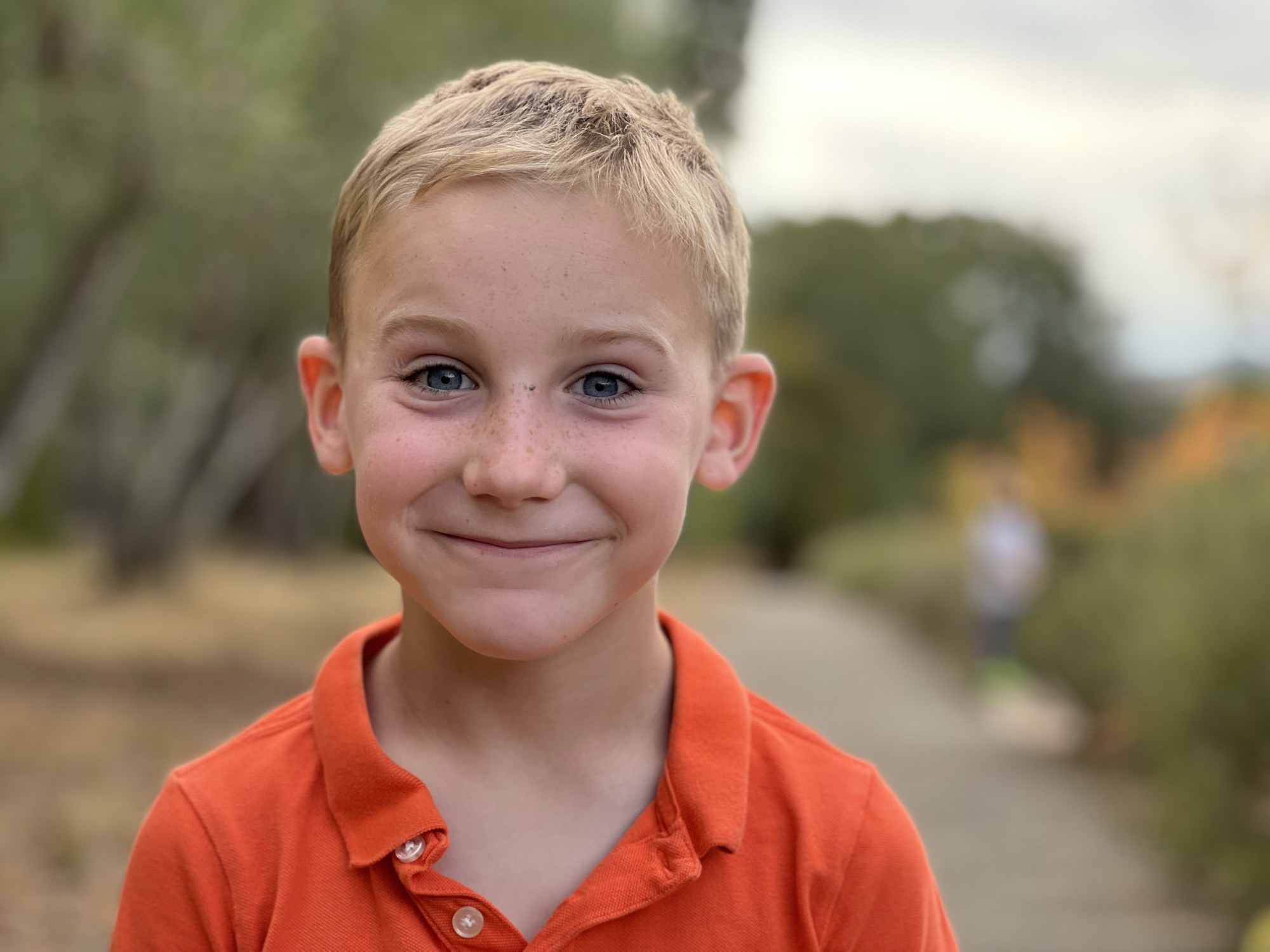 A close-up portrait of a smiling, blonde-haired boy with blue eyes and freckles wearing an orange polo shirt. He is standing outdoors on a paved path with trees and foliage blurred in the background.