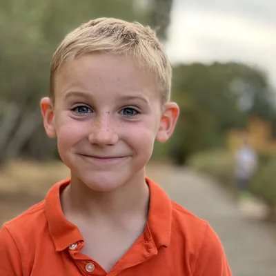A close-up portrait of a smiling, blonde-haired boy with blue eyes and freckles wearing an orange polo shirt. He is standing outdoors on a paved path with trees and foliage blurred in the background.