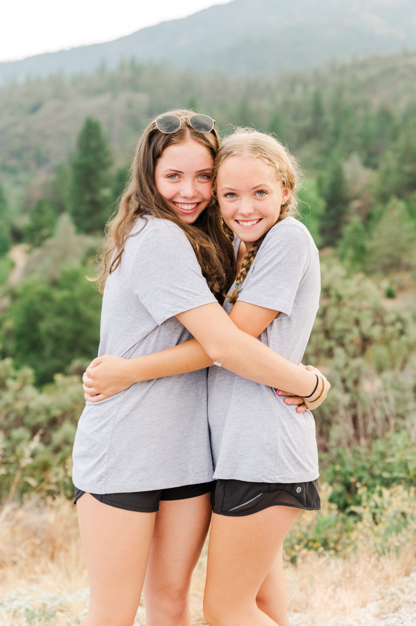 Two smiling young women in matching grey t-shirts and black shorts hug each other while posing for a photo. They are standing outdoors in a mountainous, forested area.