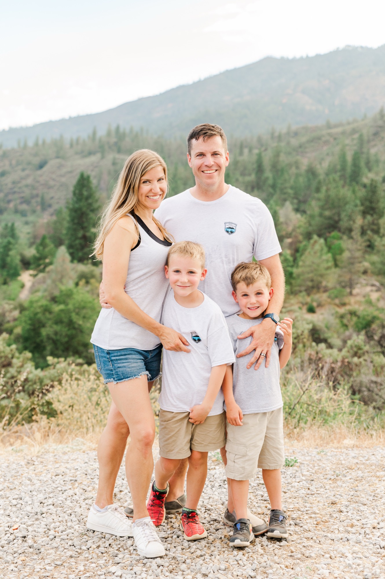 A family of four, consisting of a man, a woman, and two young boys, stands together smiling for a portrait on a gravel path. They are posed outdoors with a scenic background of forested hills and mountains.