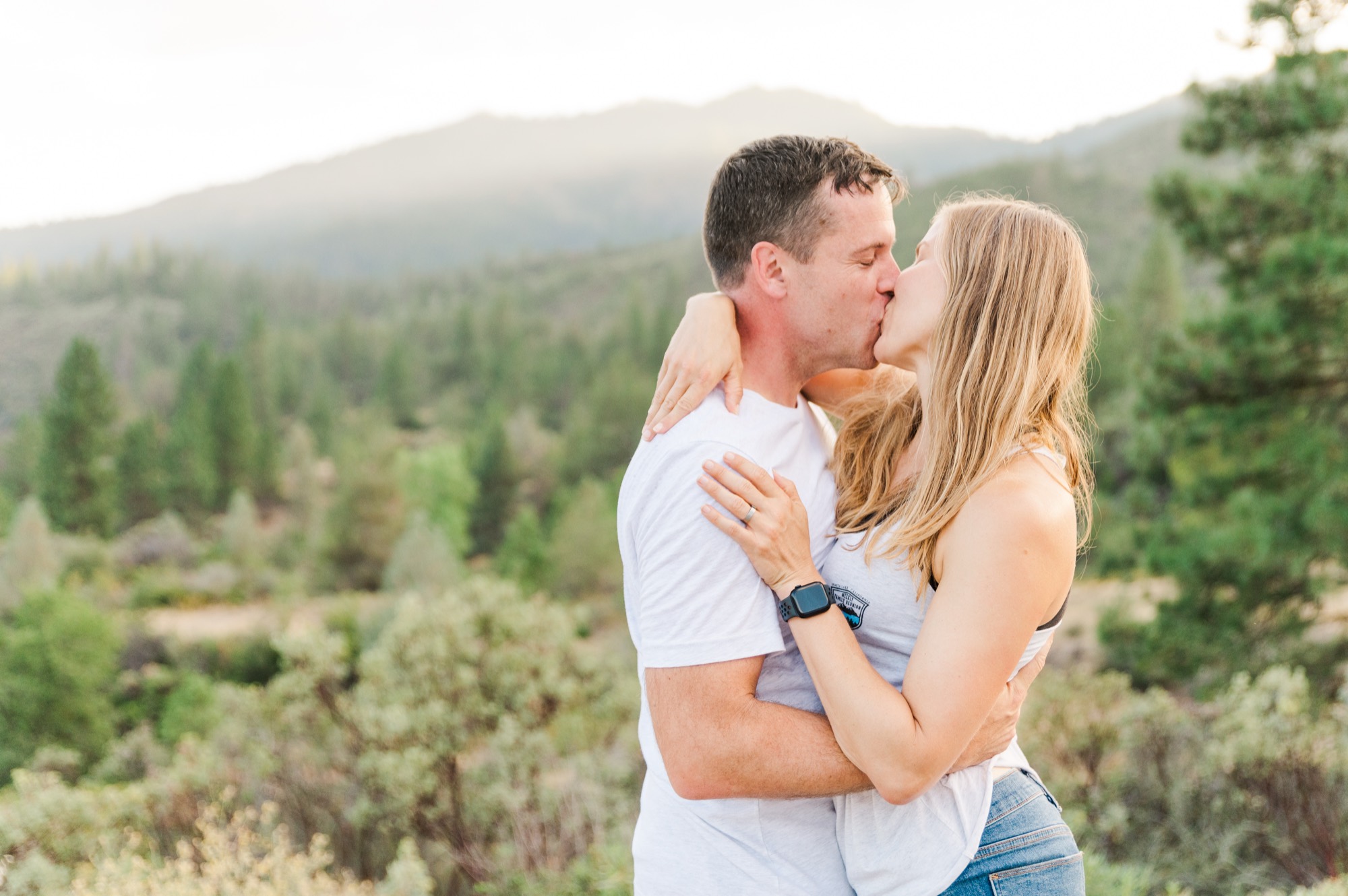 A man and a woman are embracing and kissing outdoors in a mountainous, forested landscape. They are both wearing casual clothing, with the woman's hand resting on the man's chest, showing a wedding band and a smartwatch.