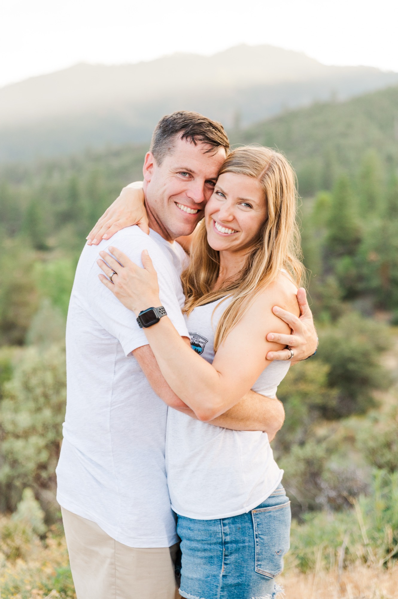 A smiling man and woman embrace outdoors, posing for a portrait with a scenic, mountainous landscape in the background.