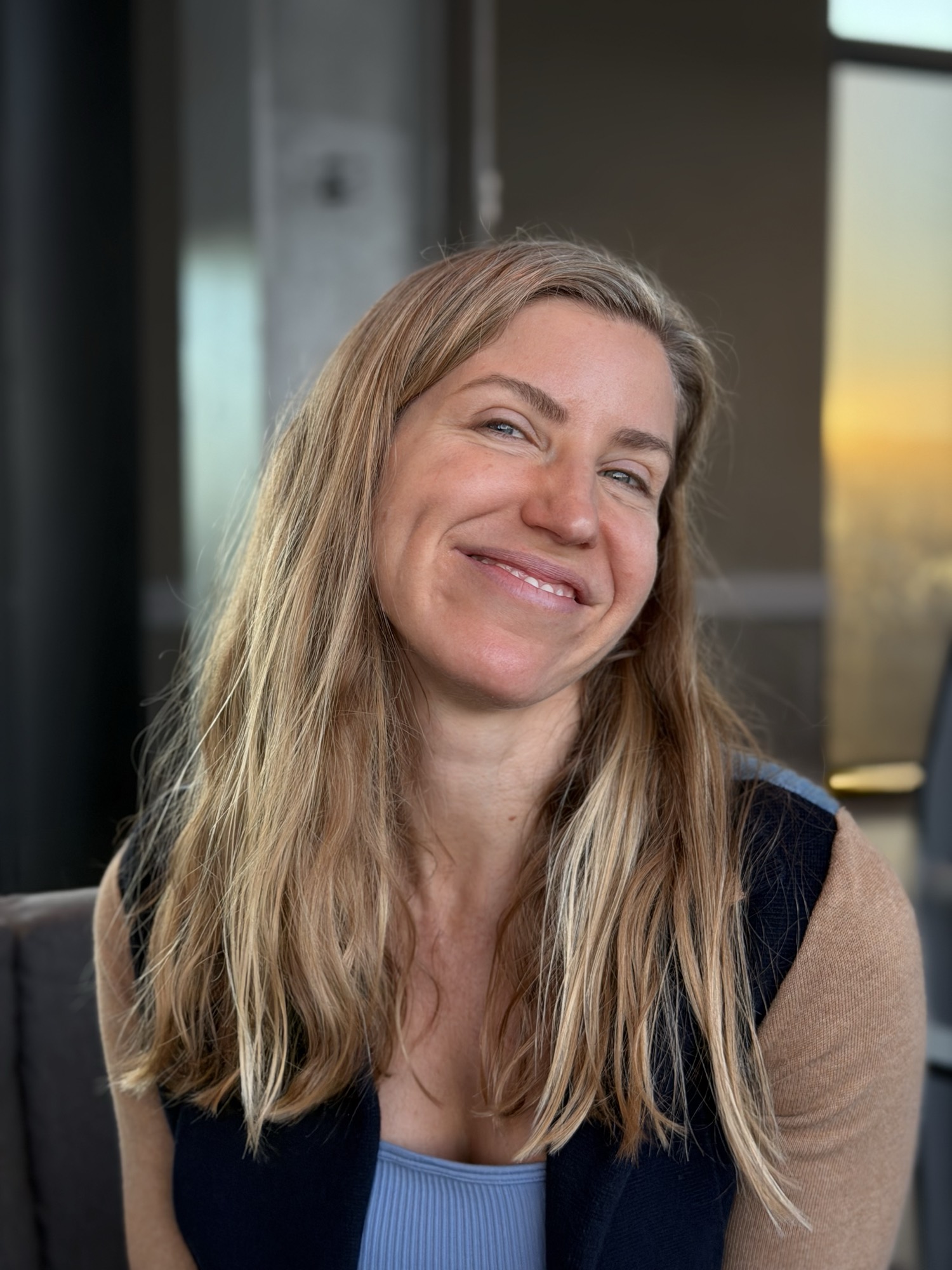 A close-up portrait shows a smiling woman with long blonde hair tilting her head slightly while posing indoors. She is wearing a blue top under a dark vest and a tan cardigan, with a softly blurred background showing a window with warm light.