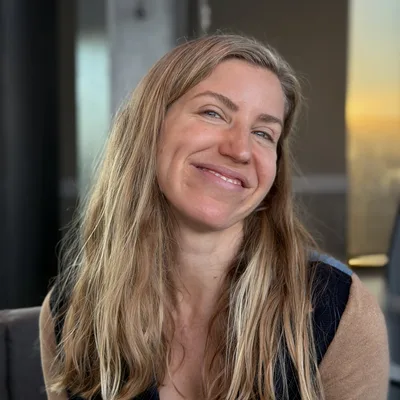 A close-up portrait shows a smiling woman with long blonde hair tilting her head slightly while posing indoors. She is wearing a blue top under a dark vest and a tan cardigan, with a softly blurred background showing a window with warm light.