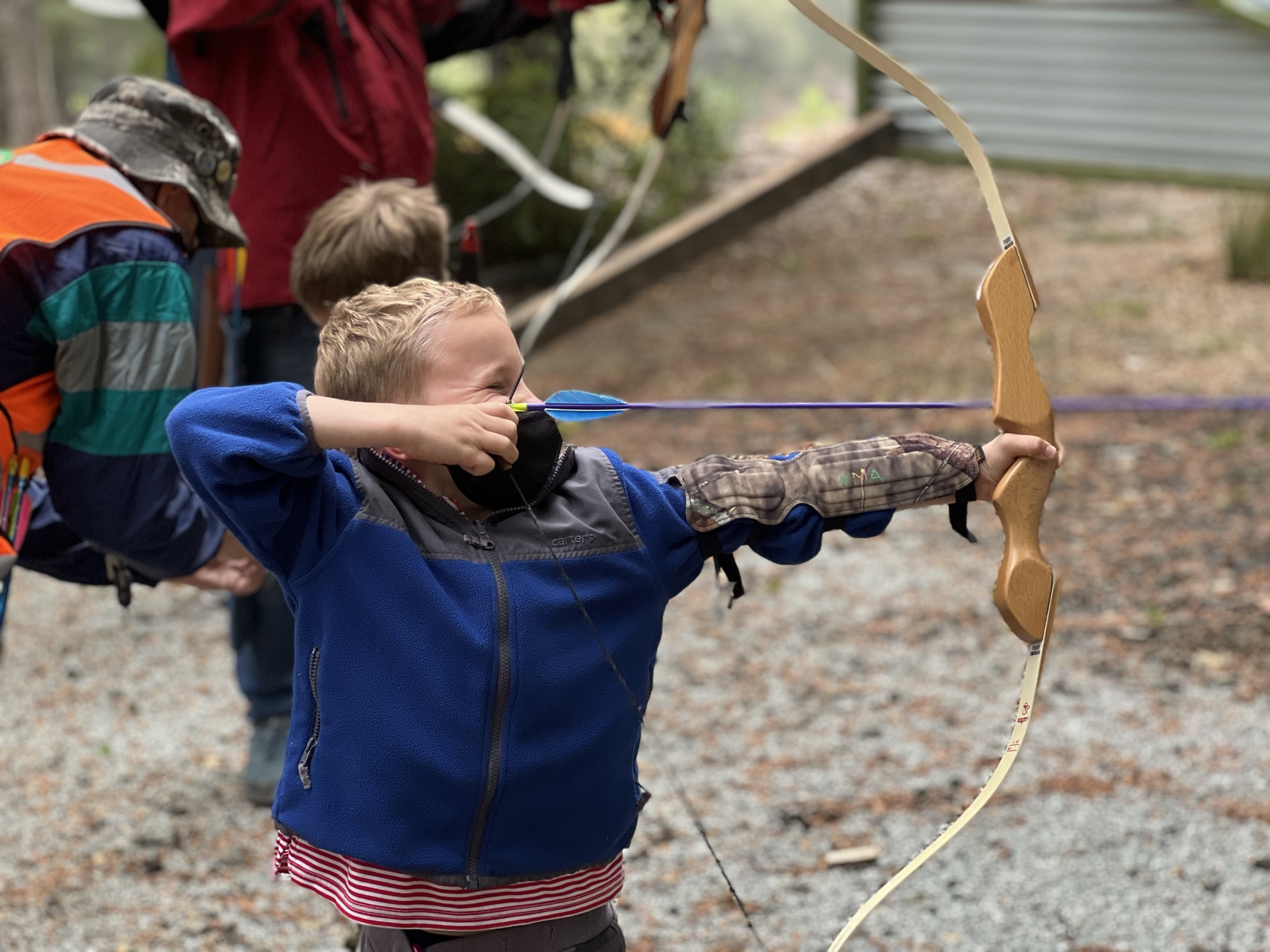 A young boy in a blue fleece jacket and arm guard aims a bow and arrow at an outdoor archery range. Other people, including an instructor in a safety vest, stand nearby on the gravel ground.