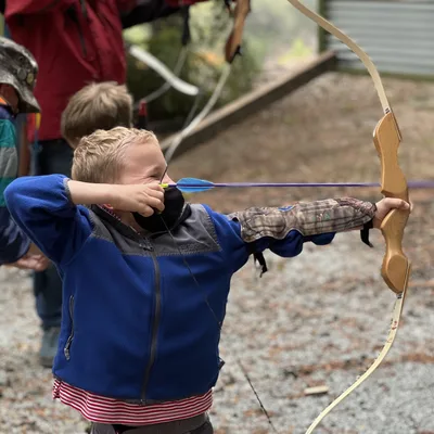 A young boy in a blue fleece jacket and arm guard aims a bow and arrow at an outdoor archery range. Other people, including an instructor in a safety vest, stand nearby on the gravel ground.