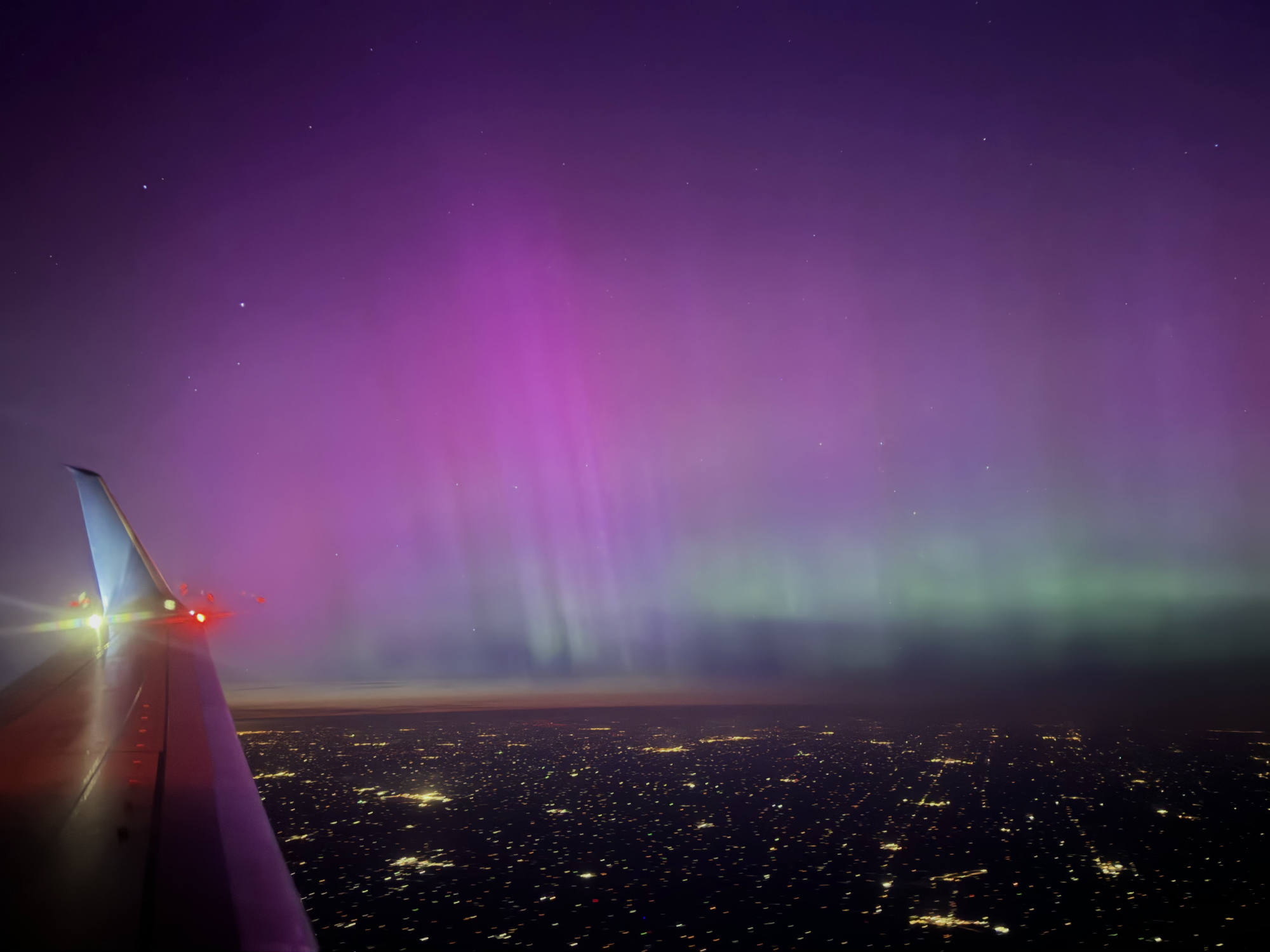 An aerial view from an airplane window captures vibrant purple and green aurora borealis lights stretching across the night sky. Below, the wing of the plane is visible on the left, overlooking a vast city grid illuminated by streetlights.