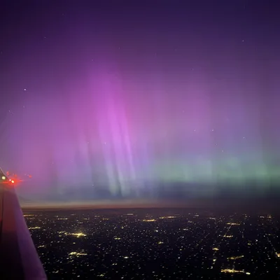 An aerial view from an airplane window captures vibrant purple and green aurora borealis lights stretching across the night sky. Below, the wing of the plane is visible on the left, overlooking a vast city grid illuminated by streetlights.