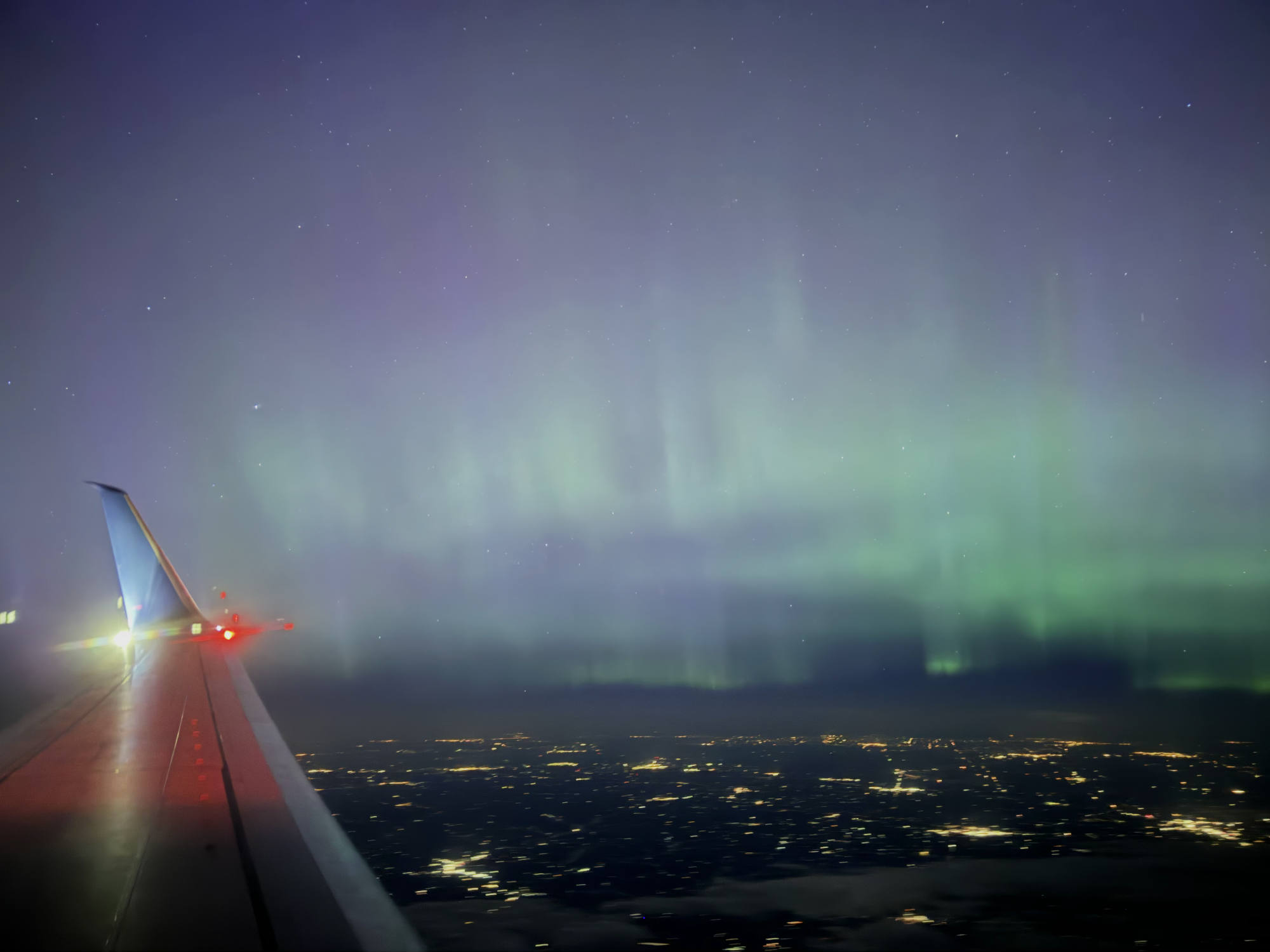 An aerial view from an airplane window captures the green and purple aurora borealis glowing in the night sky above a city's distant lights. The wing of the airplane is visible in the lower-left corner of the frame.