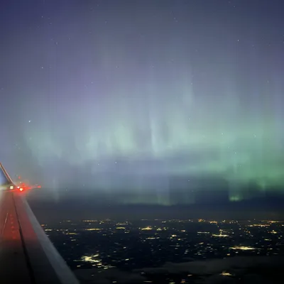 An aerial view from an airplane window captures the green and purple aurora borealis glowing in the night sky above a city's distant lights. The wing of the airplane is visible in the lower-left corner of the frame.