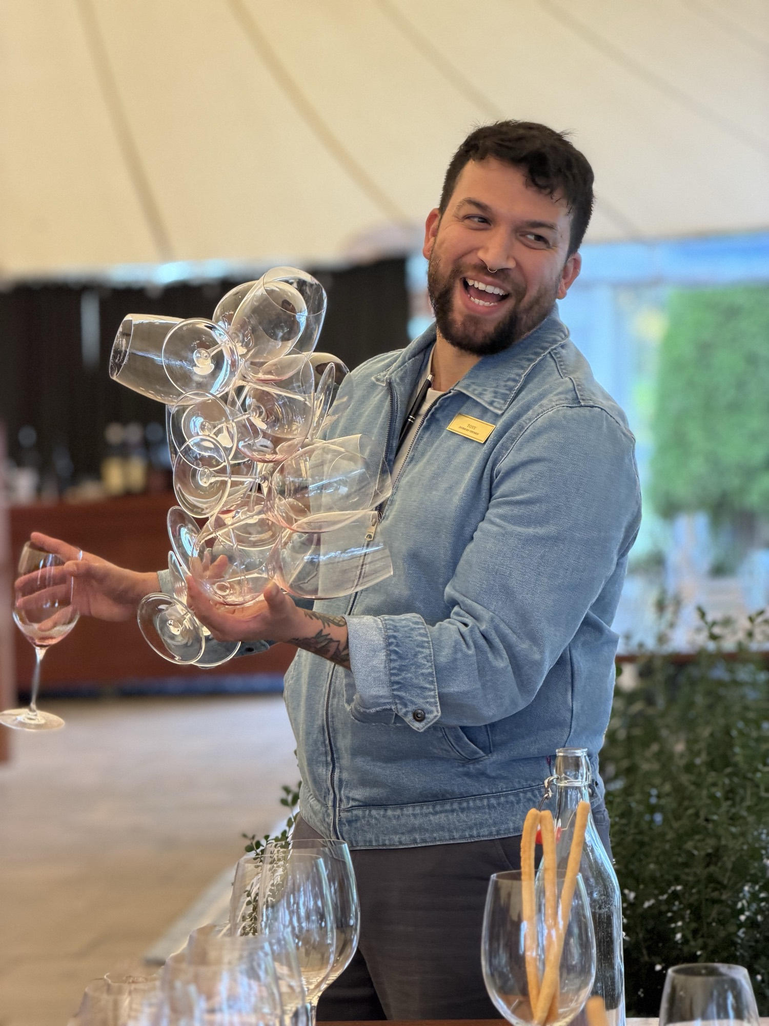A smiling man in a denim jacket stands in an outdoor event tent, holding a large, stacked cluster of empty wine glasses in one hand and a single wine glass in the other. He is positioned behind a table set with additional glassware, breadsticks, and a bottle of water.