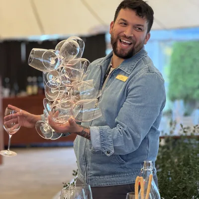A smiling man in a denim jacket stands in an outdoor event tent, holding a large, stacked cluster of empty wine glasses in one hand and a single wine glass in the other. He is positioned behind a table set with additional glassware, breadsticks, and a bottle of water.