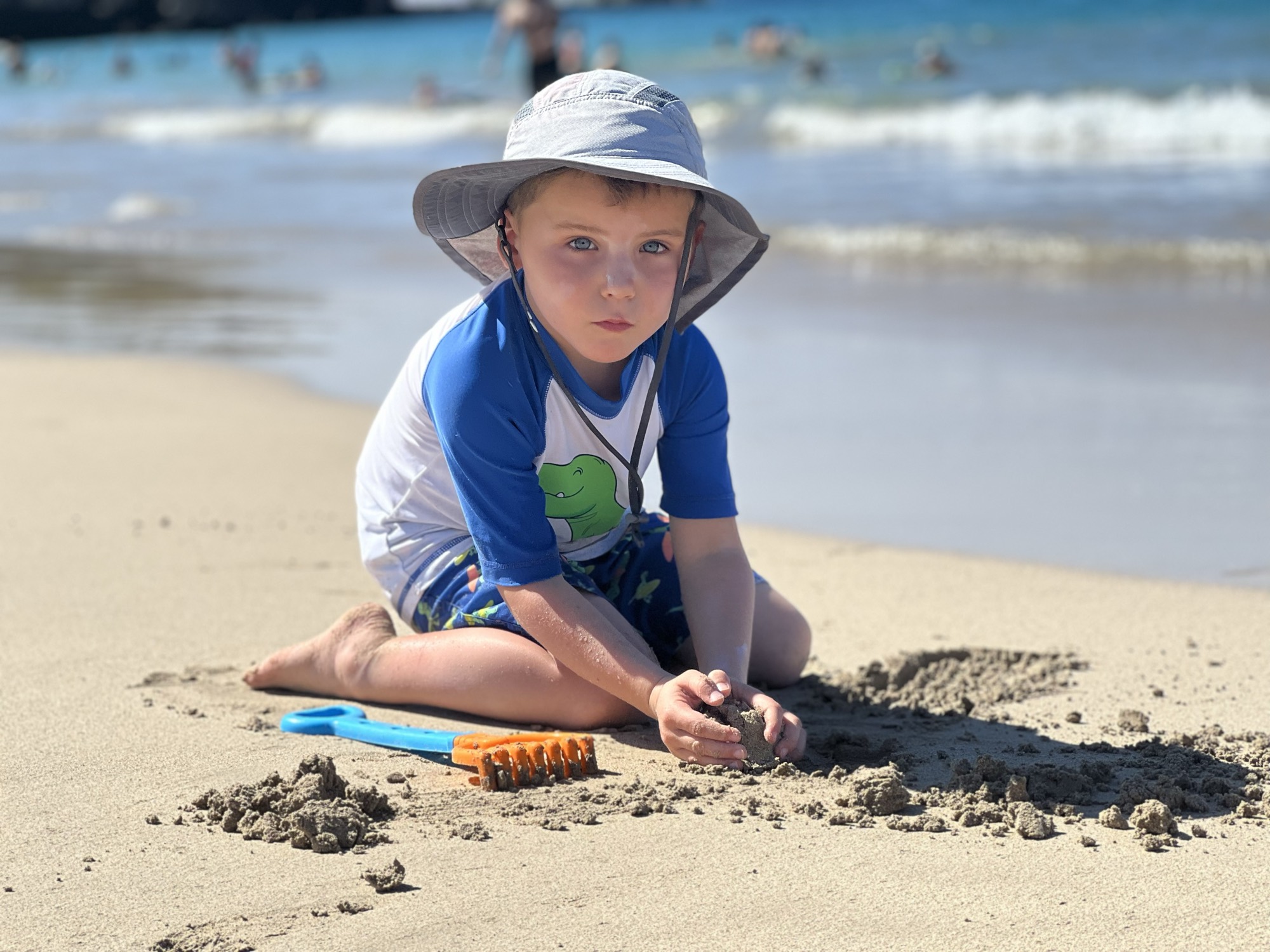 A young boy wearing a sun hat and a rash guard sits on a sandy beach, playing with wet sand near a blue and orange toy rake. In the background, waves roll onto the shore of a sunny beach with other people in the distance.