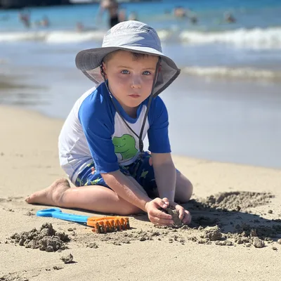 A young boy wearing a sun hat and a rash guard sits on a sandy beach, playing with wet sand near a blue and orange toy rake. In the background, waves roll onto the shore of a sunny beach with other people in the distance.